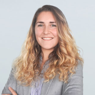 Lächelnde Person mit langen lockigen Haaren, die ein gestreiftes Hemd und einen grauen Blazer vor einem schlichten Hintergrund trägt.