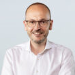 A smiling person with glasses and a beard is wearing a white shirt against a plain background.