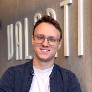 Person with glasses and short hair smiling, standing in front of a wall with large, partially visible text.