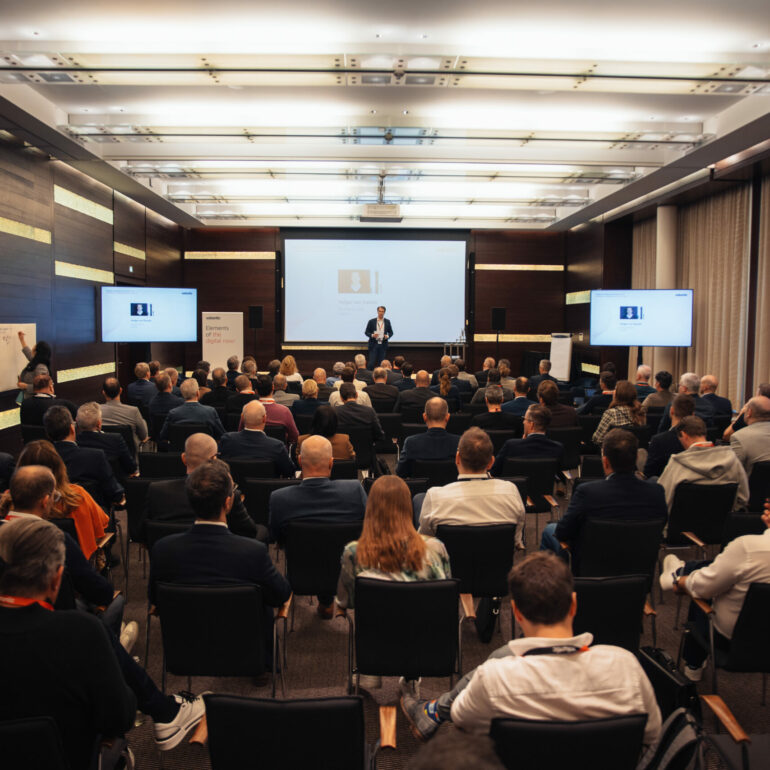 A speaker presents on stage to an audience in a conference room, with screens displaying slides and attendees seated and listening.
