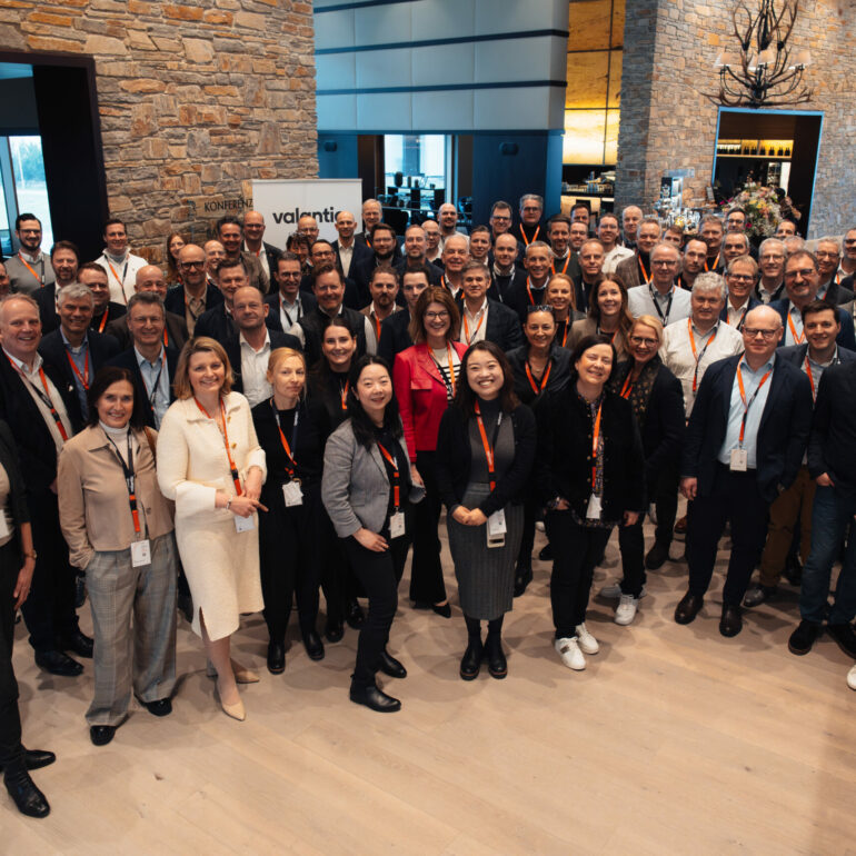 A large group of people, dressed in business attire, gather in a spacious, modern indoor setting with stone walls and wooden floors, smiling and posing for a group photo at the DEF25 conference celebrating digital excellence.