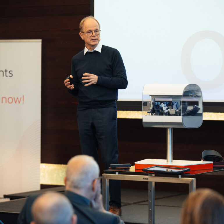 A man in a dark sweater presents at a conference, standing beside a small transparent machine on a table. A banner with "Elements of the digital now!" is visible. Audience members watch.