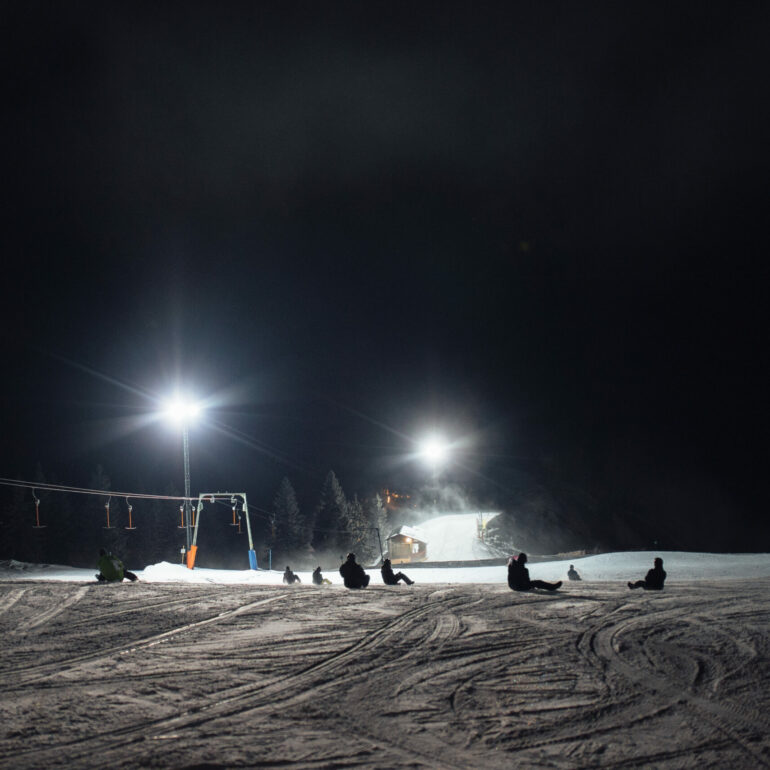 A group of people sit on a snowy slope at night, under bright lights and next to a ski lift. Trees are visible in the background.
