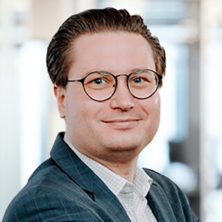 At Hannover Messe, a smiling person in glasses and a suit jacket poses confidently in a bright indoor setting.