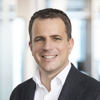 A person with short hair, wearing a white shirt and dark suit jacket, smiling at the camera in an office setting that echoes the innovative spirit of Hannover Messe.
