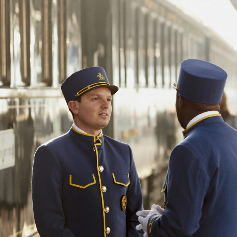 Zwei railtour-Zugbegleiter in blauen Uniformen mit goldenen Details unterhalten sich auf einem Bahnsteig vor einem historischen Zug. Im Hintergrund sind weitere Reisende zu sehen.
