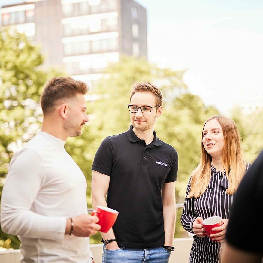 Three people standing outdoors, holding red mugs and conversing, with trees and a building in the background.