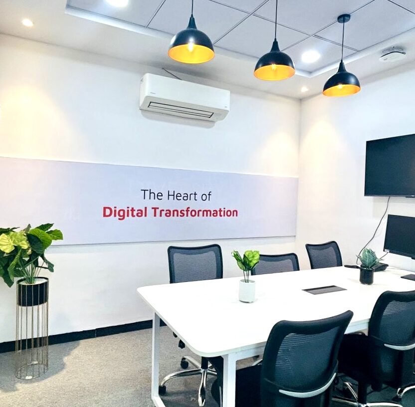 Modern conference room with a white table, black chairs, and a wall-mounted screen that embodies "The Heart of Digital Transformation." Three hanging pendant lights and plants bring an air of Indore’s vibrant charm to the space.