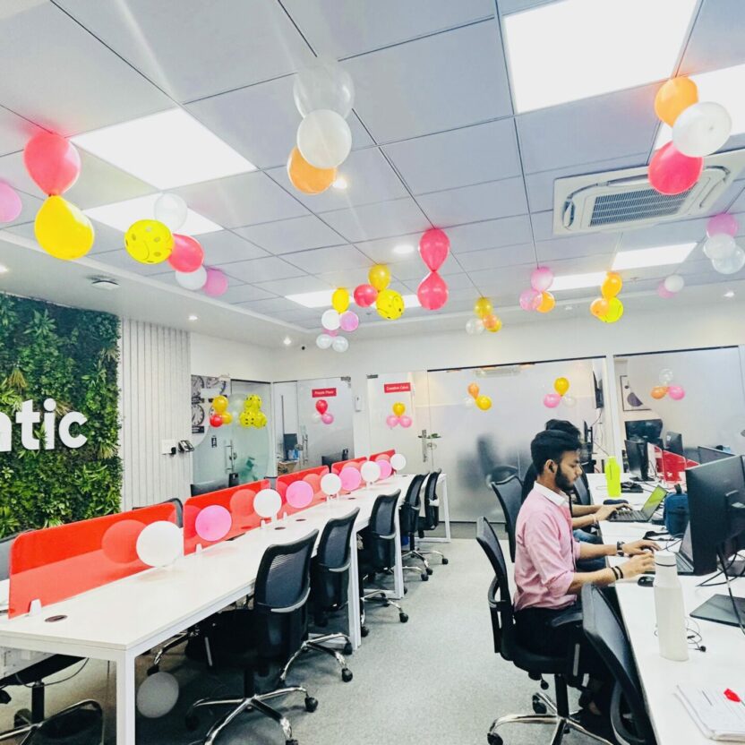 A modern office in Indore features a lush green plant wall, colorful balloons on the ceiling, vibrant red and white partitioned desks, and a person diligently working at a computer.