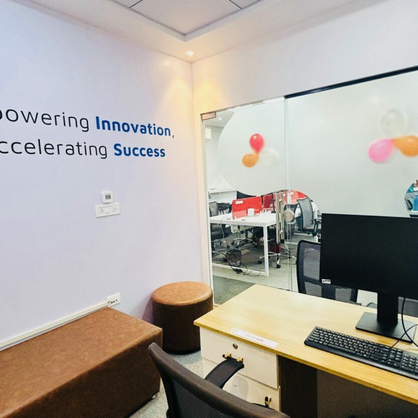 Office space in Indore features a desk, computer, and chair. A wall displays the message, "Empowering Innovation, Accelerating Success," while a glass window reveals a room festively decorated with balloons.