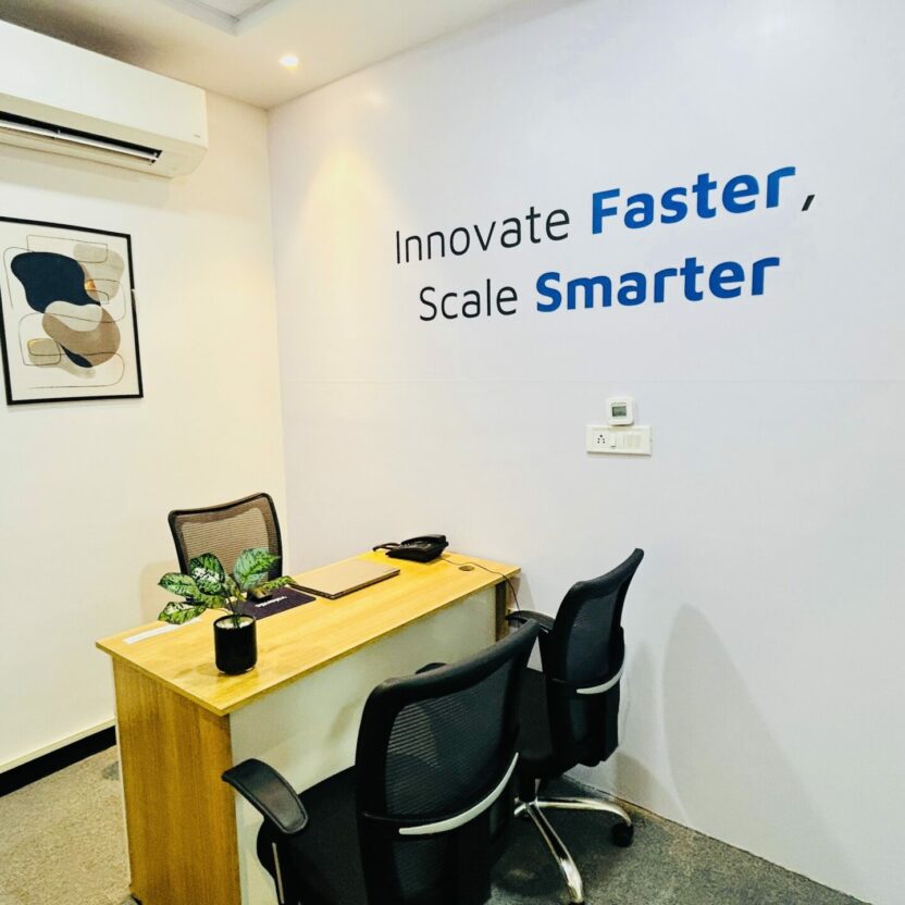 A small office in Indore features a wooden desk, two chairs, and potted plants. A wall sign reads "Innovate Faster, Scale Smarter," beneath an air conditioner that hangs above framed abstract artwork.