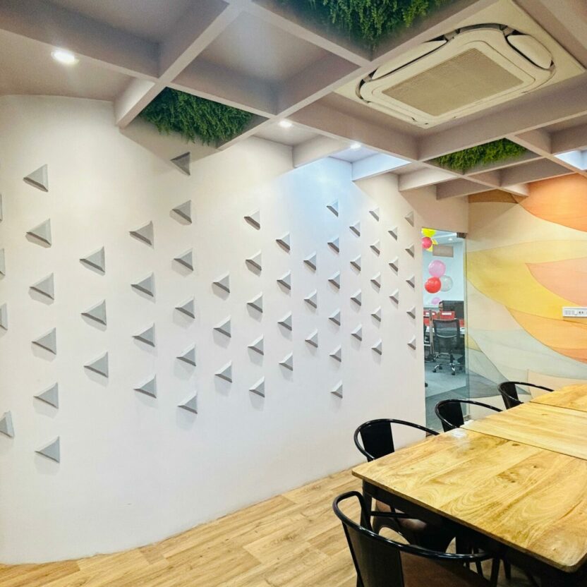 Modern meeting room in Indore featuring a long wooden table and sleek black chairs. The space is highlighted by a curved wall with geometric patterns, a ceiling adorned with green accents, and a vibrant mural. Equipped with air conditioning and cameras for convenience.