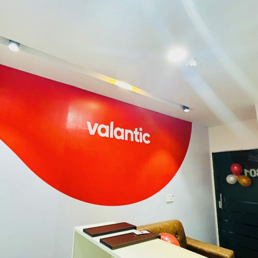 The reception area features a striking curved red wall adorned with the "Valantic" logo. Three clocks display times from different cities, including Indore. Ceiling lights add brightness, while an air conditioner ensures comfort.