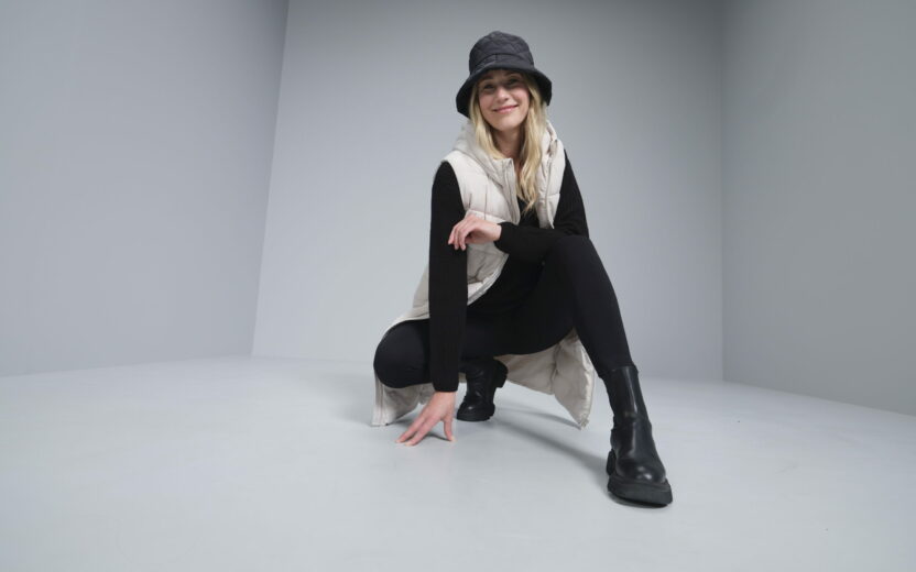 TAKKO Fashion Omnichannel: Person in black outfit with white vest and bucket hat squatting in a gray room, smiling.