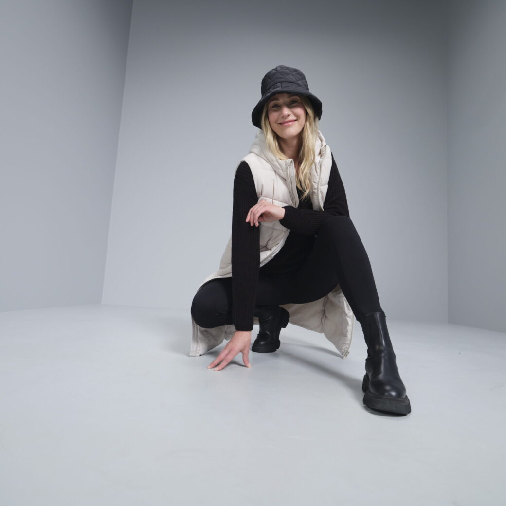 TAKKO Fashion Omnichannel: Person in schwarzem Outfit mit weißer Weste und Anglerhut hockt in grauem Raum und lächelt.