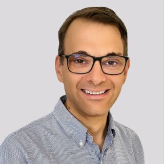 Eine lächelnde Person mit Brille und hellblauem Hemd steht vor einem schlichten grauen Hintergrund und verkörpert den Geist der vernetzten Planung.