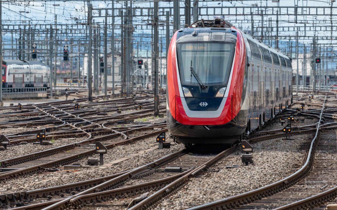 A modern train travels on a railway track surrounded by complex overhead wiring and multiple tracks at a rail yard.