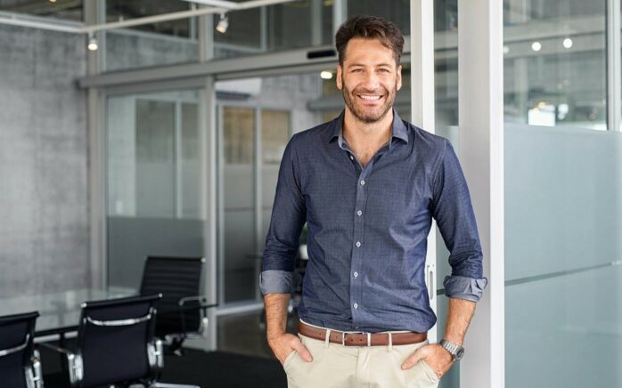 Ein lächelnder Mann steht in einem modernen Büroraum mit Glaswänden und schwarzen Stühlen im Hintergrund, was die nahtlose Integration widerspiegelt, die SAP S/4HANA Unternehmen bietet.