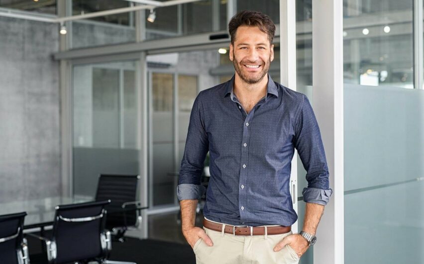 Ein lächelnder Mann steht in einem modernen Büroraum mit Glaswänden und schwarzen Stühlen im Hintergrund, was die nahtlose Integration widerspiegelt, die SAP S/4HANA Unternehmen bietet.