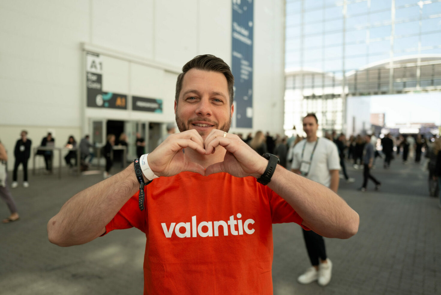 A man in an orange "valantic" shirt makes a heart shape with his hands outside a convention center, with people and event signs in the background.