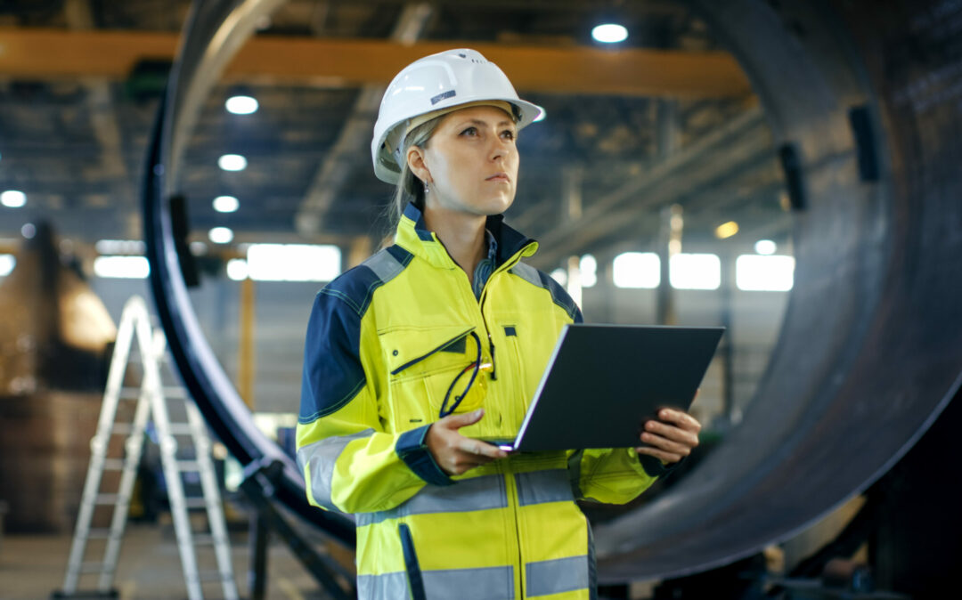 Female Industrial Engineer in the Hard Hat Uses Laptop Computer while Standing in the Heavy Industry Manufacturing Factory. In the Background Various Metalwork Project Parts Lying