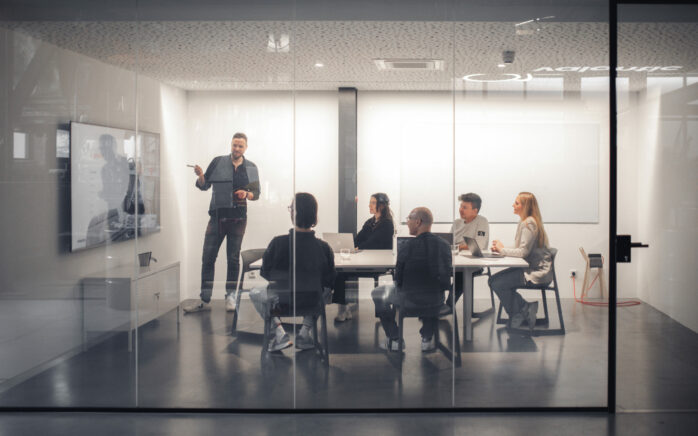Eine Gruppe von sechs Personen hält sich in einem modernen Konferenzraum mit Glaswänden auf: Während ein Mann vorne steht und eine Präsentation auf einem großen Bildschirm hält, sitzen die anderen an einem Konferenztisch und hören zu. Einige haben Laptops vor sich. Der Raum ist hell beleuchtet und minimalistisch eingerichtet.