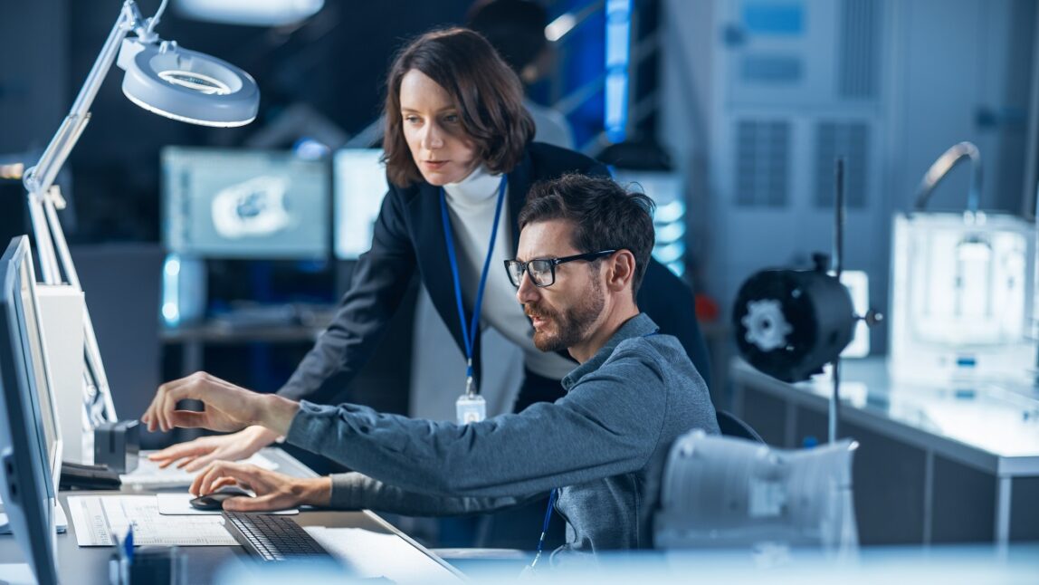 Futuristic Machine Engine Development Engineer Working on Computer at His Desk, Talks with Female Project Manager. Team of Professionals Working in the Modern Industrial Design Institution, discussing SAP APM.