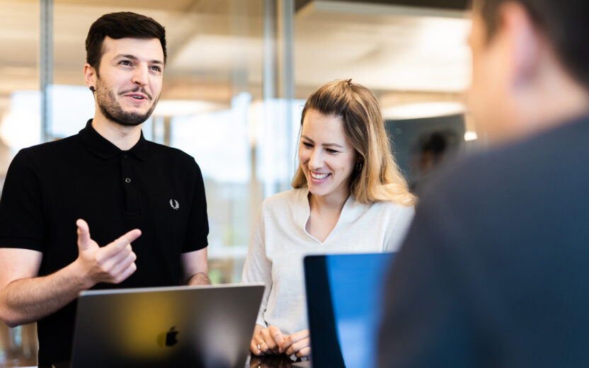 Drei Personen stehen in einer modernen Büroumgebung und unterhalten sich über die Einführung von SAP S/4HANA. Eine Person im schwarzen Polohemd gestikuliert,