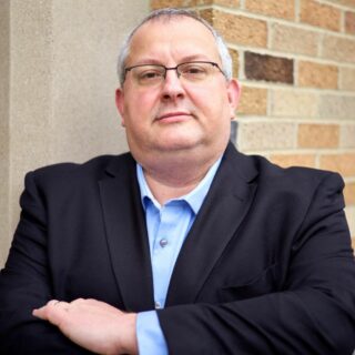 Ein Mann mittleren Alters in schwarzer Anzugsjacke und blauem Hemd steht mit verschränkten Armen vor einer Backsteinmauer, als würde er darüber nachdenken, was wäre, wenn", und blickt nachdenklich in die Kamera.