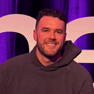 A man with short dark hair and a beard, wearing a dark hoodie, sits in front of a purple backdrop and large white letters at the Connected Manufacturing Forum 2025.