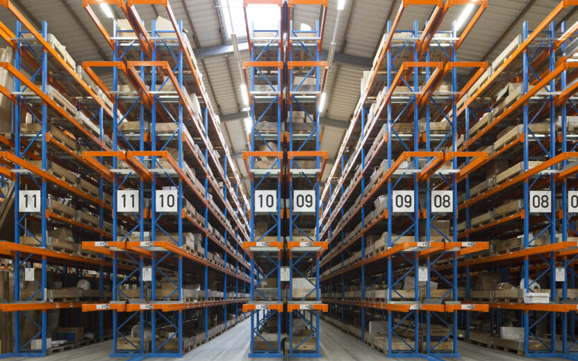 Ein großes Lager mit hohen Regalen in Blau und Orange, beschriftet mit Abschnittsnummern, gefüllt mit Kartons und Paletten. Der Raum ist hell beleuchtet und besitzt ein hohes Metalldach.