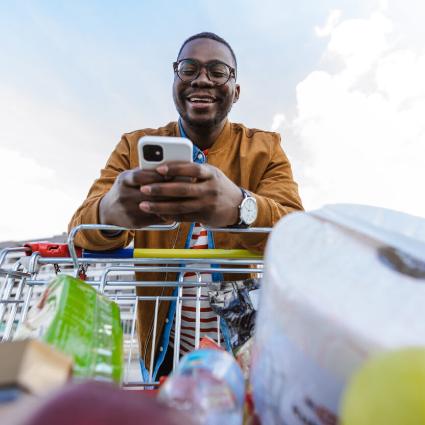 Portrait of a happy young African-American man using smart phone and pulling a shopping cart