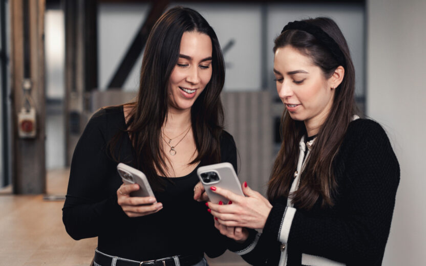 Zwei Frauen, die in einem Haus stehen, lächeln und gemeinsam auf ihre Smartphones schauen und sich möglicherweise über Social Media Marketing unterhalten.