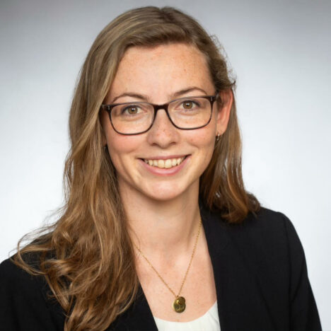 Eine Frau mit langen hellbraunen Haaren und Brille, die einen schwarzen Blazer und eine Halskette trägt, lächelt in die Kamera vor einem schlichten Hintergrund.