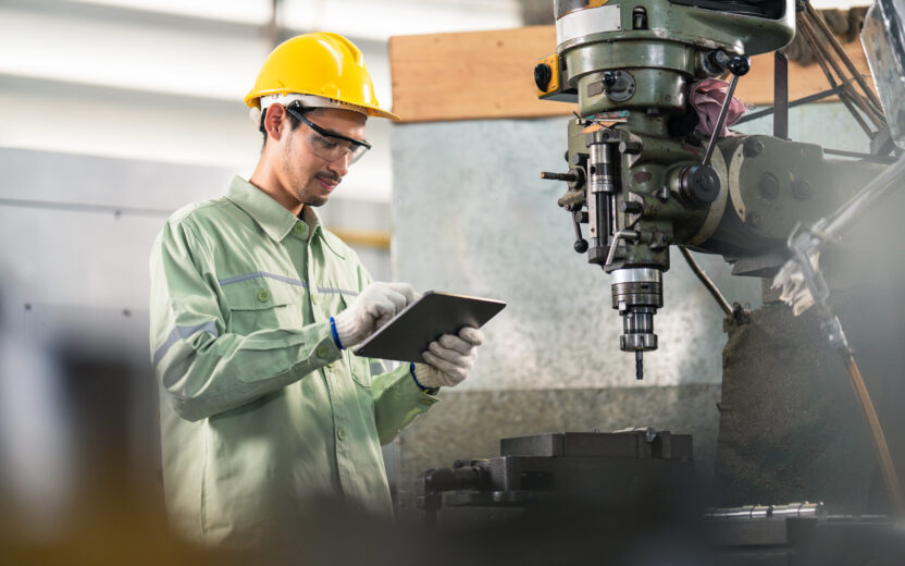 Ein Arbeiter in Schutzkleidung und gelbem Helm hält ein Tablet in den Händen und steht neben einer großen Industriemaschine in einer Fabrikumgebung.
