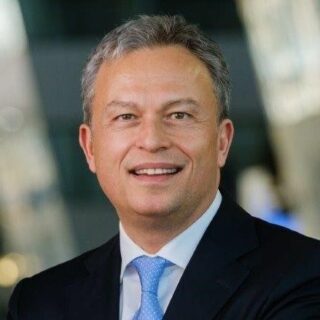 A middle-aged man in a suit and blue tie smiles at the camera, standing indoors with a blurred background at the Digital Excellence Forum.