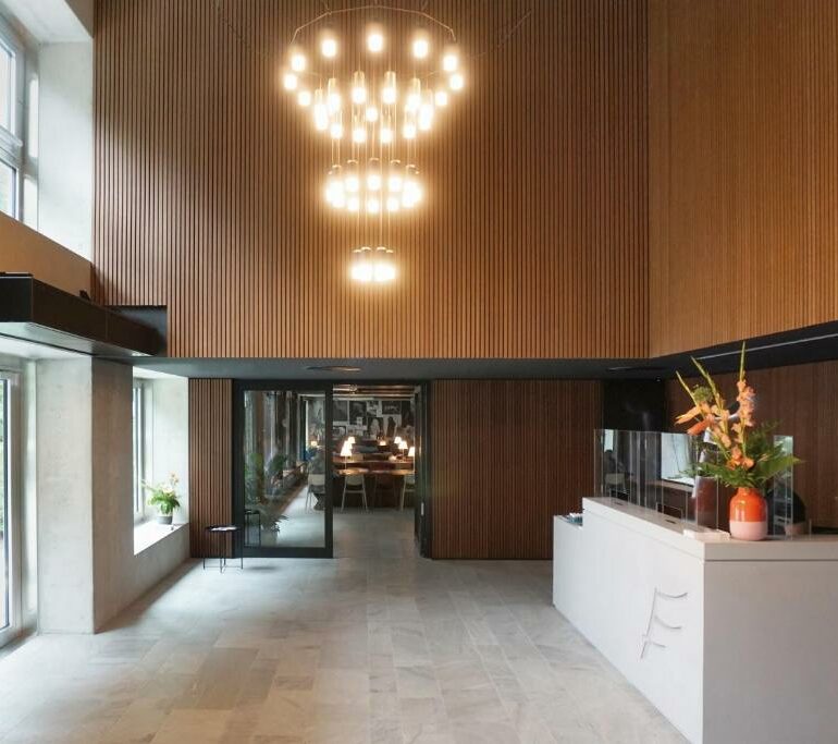 Wie wäre es, wenn diese moderne Lobby mit großen Fenstern, holzgetäfelten Wänden, einem weißen, blumengeschmückten Empfangstresen und moderner Kronleuchter-Beleuchtung Ihr neuer Lieblingsplatz zum Entspannen wäre?.