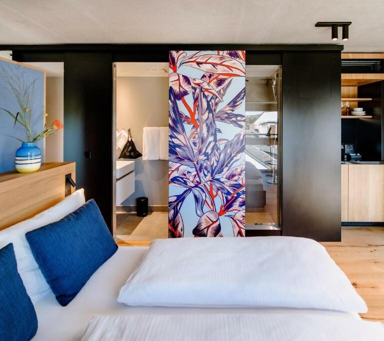 Ein modernes Hotelzimmer mit einem sauber gemachten Bett, blauen und weißen Kissen, einem dekorativen Paneel mit buntem Blattmuster und einem offenen Blick auf das Badezimmer und die Küchenzeile - wie wäre es, wenn Ihr perfekter Aufenthalt hier beginnen würde?.