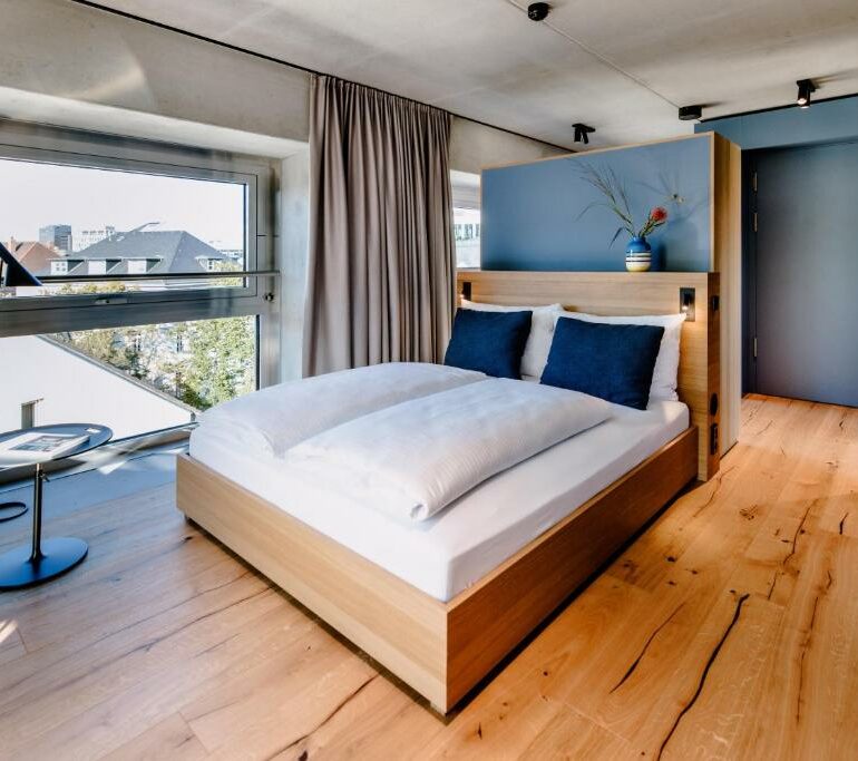 Wie wäre es, wenn Sie sich in einem modernen Schlafzimmer mit Holzboden, einem Doppelbett mit weißer Bettwäsche und blauen Kissen, einem großen Fenster, einem Fernseher auf einem Ständer und einer dekorativen Vase auf einer Trennwand hinter dem Bett entspannen könnten?.
