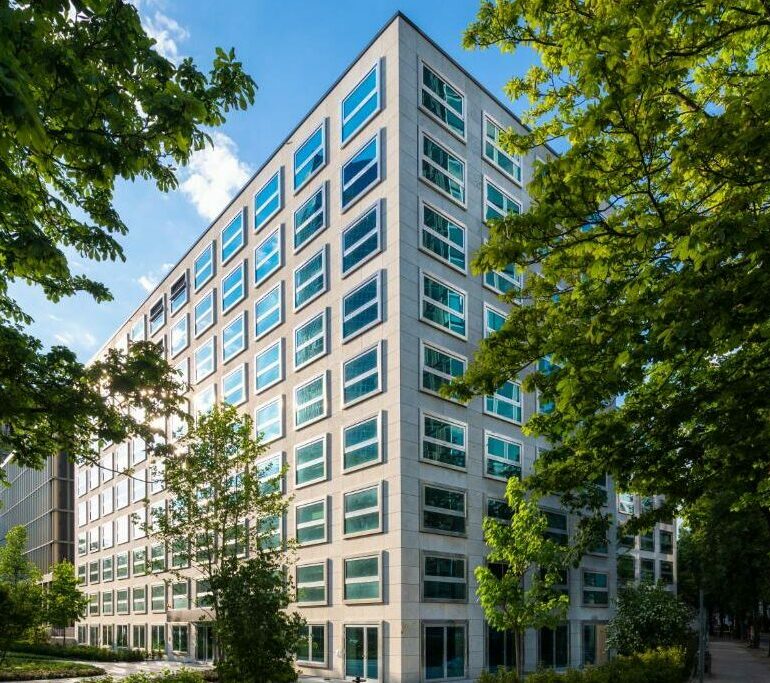 Was wäre, wenn ein modernes, weißes, mehrstöckiges Bürogebäude mit vielen Fenstern umgeben von grünen Bäumen unter einem blauen Himmel mit einigen Wolken stünde?.
