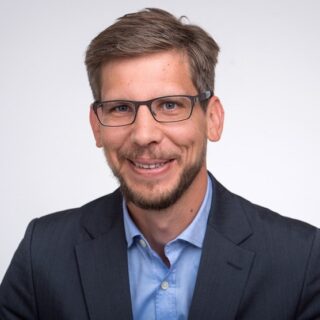 Ein Mann mit kurzen braunen Haaren, Brille und Bart trägt ein blaues Hemd und einen dunklen Blazer und lächelt vor einem schlichten, hellen Hintergrund in die Kamera, als ob er Sie dazu einladen würde, über "Was wäre wenn"-Möglichkeiten nachzudenken.