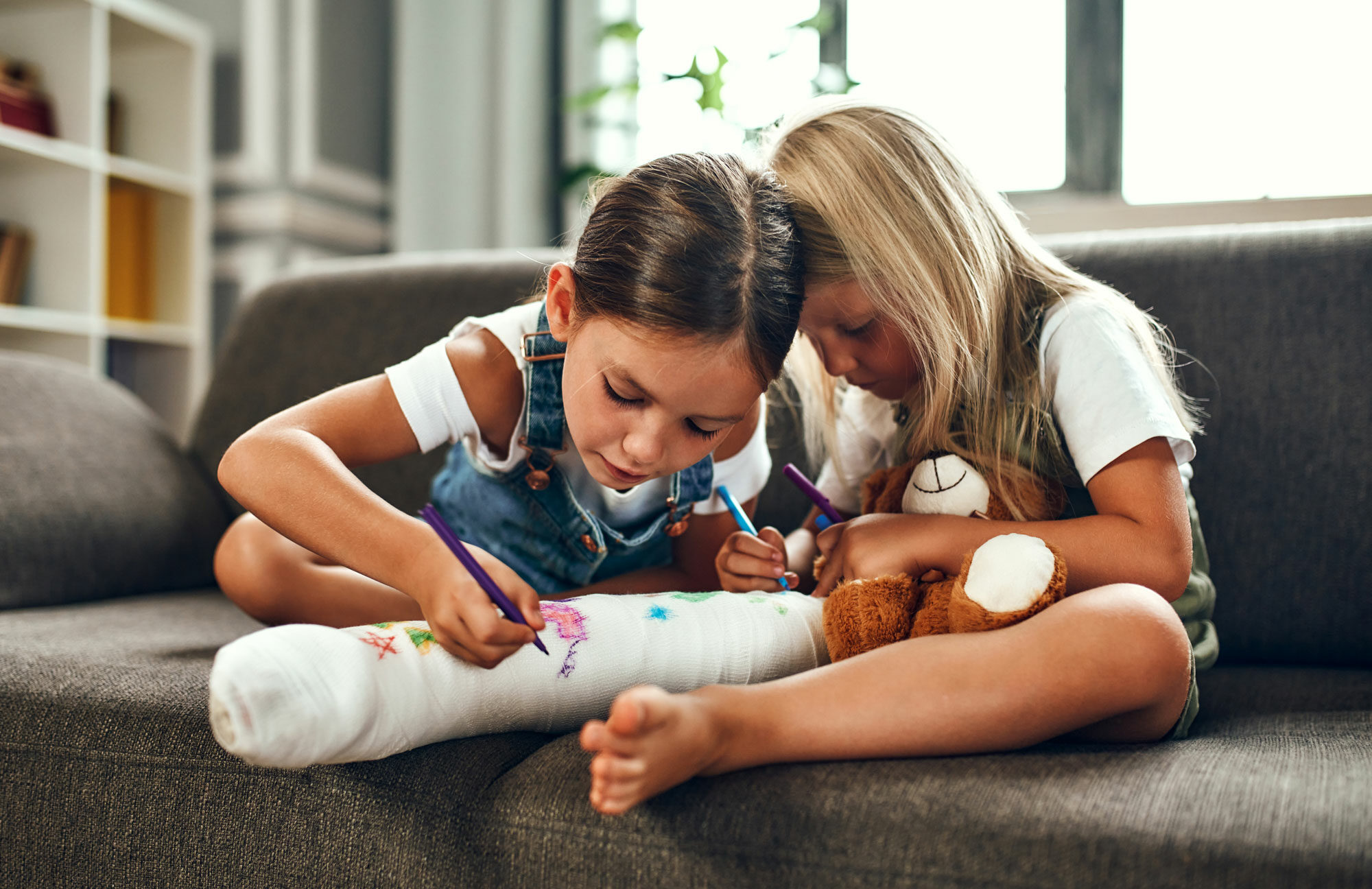 two little girls sitting on a sofa and writing on the cast on the leg of one of the girls