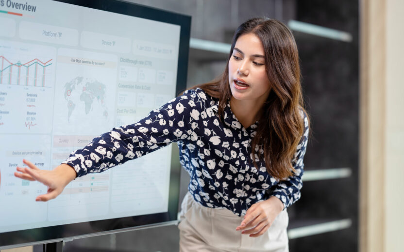 A woman in a patterned blouse points at a large digital screen displaying charts and data, illustrating the impact of digitalisation on the insurance industry during her presentation.
