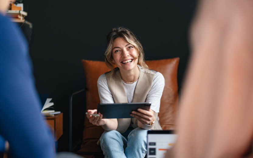 A woman sits on a brown chair, smiling and holding a tablet, engaging in conversation with two other people in an office setting about digitalisation in insurance.
