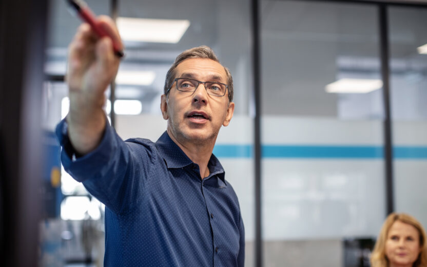 A man wearing glasses and a blue shirt points with a red pen at a board in an office, discussing digitalisation in insurance while a woman sits nearby.