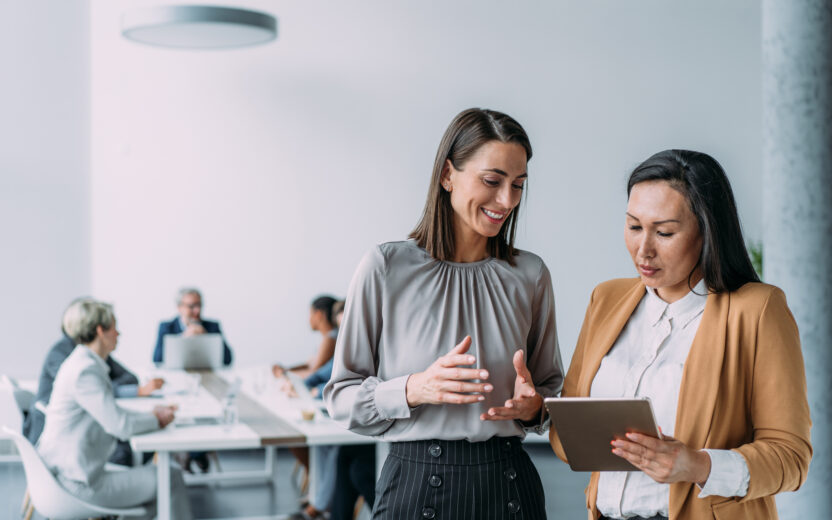 Zwei Frauen stehen und diskutieren etwas, während sie auf ein Tablet in einem modernen Büro schauen, das die Digitalisierung von Versicherungen widerspiegelt, während im Hintergrund Mitarbeiter zusammenarbeiten.