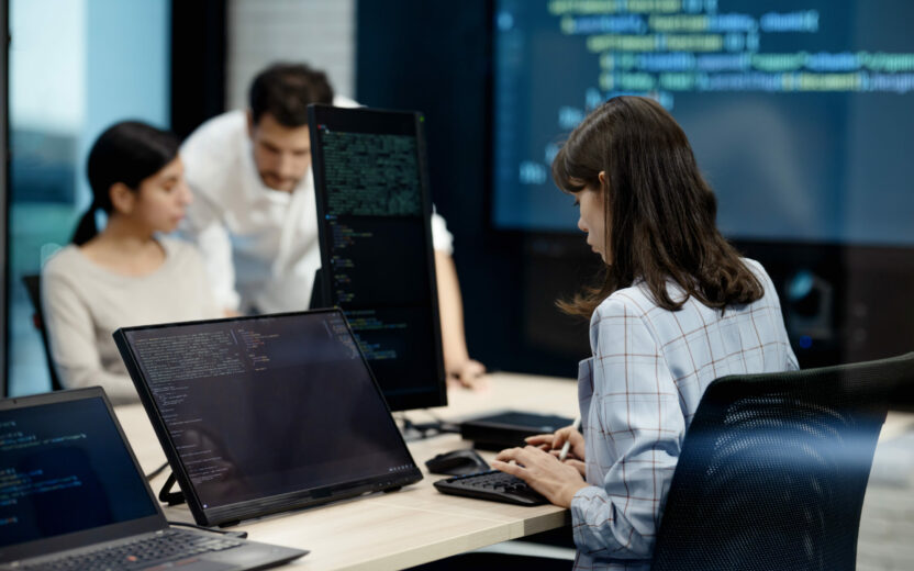 Drei Personen arbeiten an Schreibtischen mit Computern, auf denen Codes angezeigt werden, in einem modernen Büro, das sich mit Digitalisierung versicherungen beschäftigt. Eine Frau tippt an ihrem Monitor, während zwei Kollegen im Hintergrund Lösungen diskutieren.