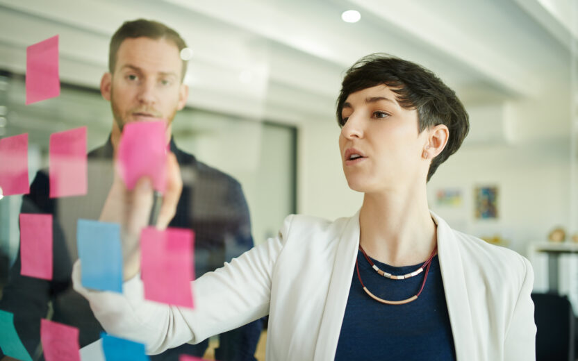 Zwei Personen in einem Büro kleben bunte Haftnotizen an eine Glaswand und scheinen gemeinsam etwas zu besprechen oder zu planen - vielleicht ein Brainstorming Ideen zur Digitalisierung von Versicherungen.