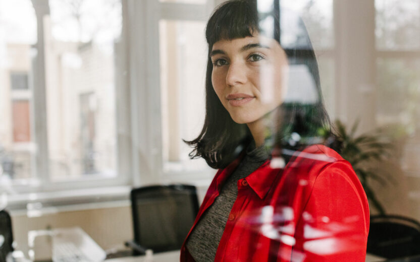 Uma mulher de camisa vermelha está em um escritório, olhando através de uma divisória de vidro com reflexos visíveis no vidro.