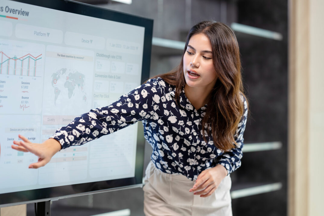 Eine Frau in einer gemusterten Bluse zeigt während einer Unternehmenspräsentation über Digitalisierung in Versicherungen auf Daten auf einem großen Bildschirm.